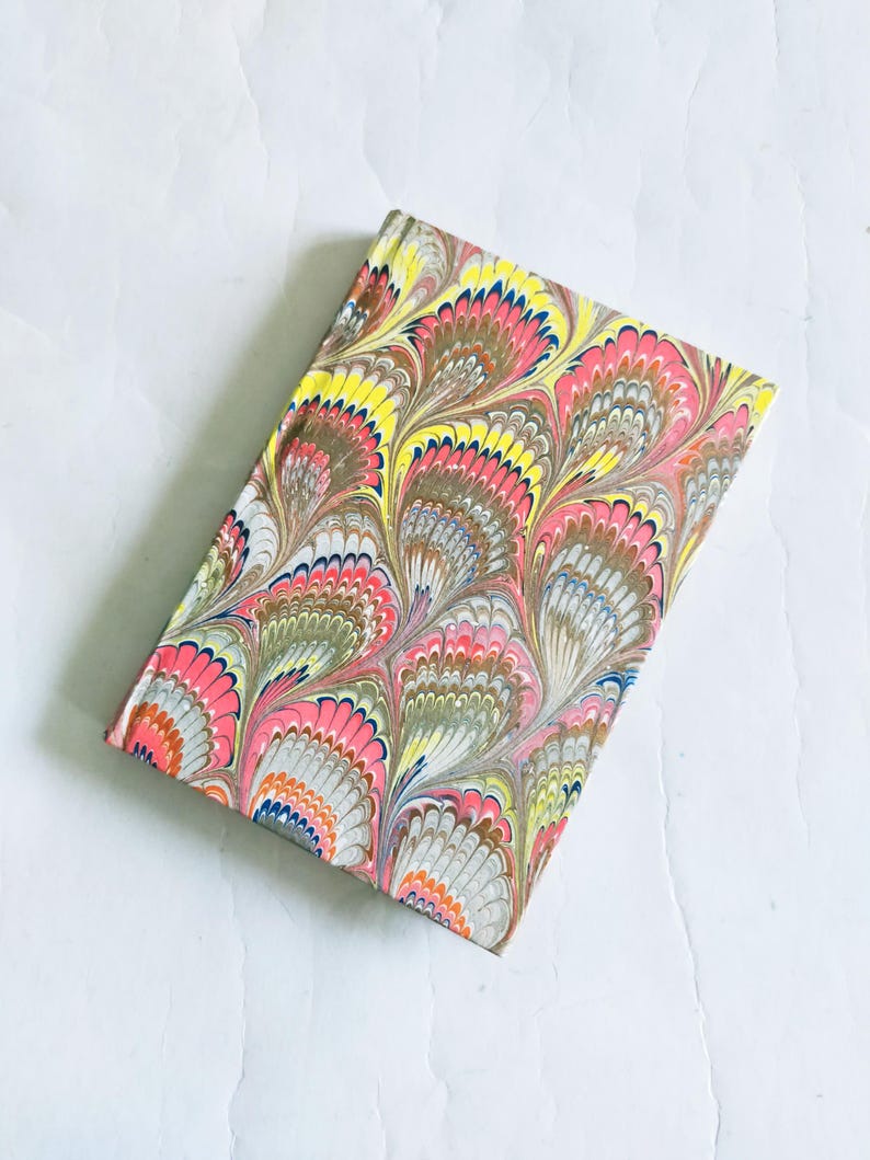 May include: A small notebook with a colorful marbled cover featuring a peacock feather design in shades of pink, yellow, blue, and brown.