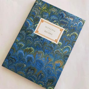 Op de afbeelding: Een receptenboek met een gemarmerd patroon in blauw, groen en goud. De omslag heeft de tekst "IN CUCINA RECIPES" in een decoratief kader. Het boek is rechthoekig en lijkt ongebruikt.