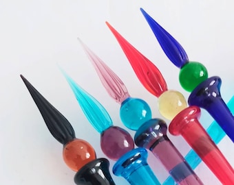 May include: A set of six colorful glass dip pens with different colored glass tips and nibs. The pens are arranged in a fan shape, with the tips pointing towards the top of the image. The pens are all different colors, including black, red, blue, green, purple, and turquoise.