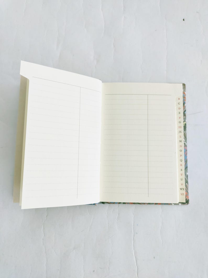 May include: An open notebook with lined pages and a colorful marbled cover. The cover has a small index with letters of the alphabet along the edge.