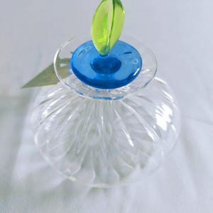 May include: A clear glass perfume bottle with a blue and green stopper. The stopper is a blue circle with a green leaf-shaped top. The bottle has a ribbed design and sits on a white surface.