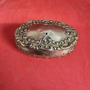 May include: An oval, silver-coloured antique box with ornate floral and scrollwork detailing. The box has a hinged lid and sits on a red surface. The box is likely made of metal and is a decorative item.