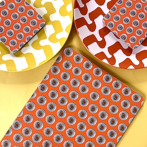 Puede incluir: Tres posavasos con estampado naranja y blanco con un diseño geométrico. Los posavasos están sobre una superficie amarilla.