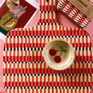 May include: A red, white, and black patterned cutting board with a matching small bowl featuring a cherry design. A set of coasters and a decorative card are also visible.