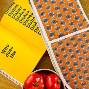 May include: A bright yellow cookbook with the text "Who does the Dishes" next to an orange oven glove with a white trim and a floral pattern. A bowl of red tomatoes is also in the image.
