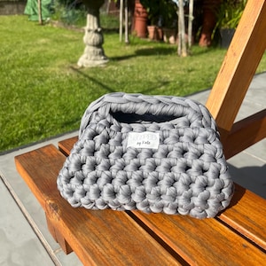May include: A dark grey handbag crafted from thick, chunky crocheted yarn. The bag features a structured, rectangular shape with a top opening and a white fabric label on the front that displays the text "LOOPED by Kate" in black lettering.