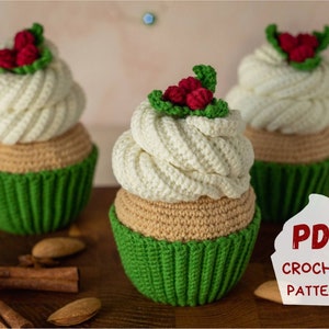 Op de afbeelding: Drie gehaakte kerstcupcake-versieringen met groene glazuur en rode hulstbesjes. De versieringen liggen op een houten oppervlak met kaneelstokjes en amandelen. Een wit bord met de tekst "PDF CROCHET PATTERN" staat op de voorgrond.