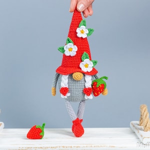 Op de afbeelding: Een gehaakte gnome met een rode muts en witte bloemen, die een rode aardbei vasthoudt. De gnome staat op een wit oppervlak met een kleine rode aardbei ernaast.