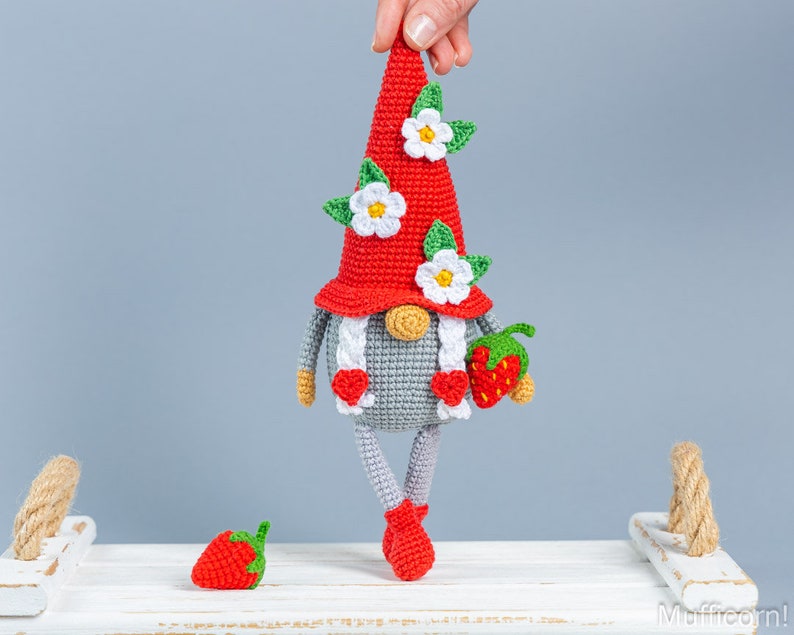 Op de afbeelding: Een gehaakte gnome met een rode muts en witte bloemen, die een rode aardbei vasthoudt. De gnome staat op een wit oppervlak met een kleine rode aardbei ernaast.