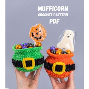 May include: Two crocheted pots in green and orange, each featuring a black band with a yellow buckle. The pots are filled with colorful candies and topped with a pumpkin-shaped lollipop and a ghost-shaped treat. Text reads MUFFICORN CROCHET PATTERN PDF.