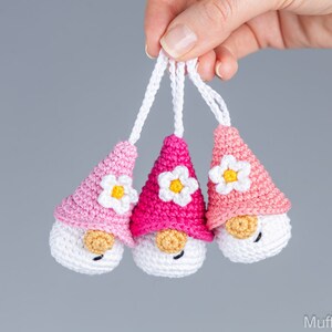 May include: Three handmade crochet gnome ornaments in shades of pink and white. Each gnome has a white face, a tan nose, and a flower on its pointed hat. The ornaments are suspended by white cords.