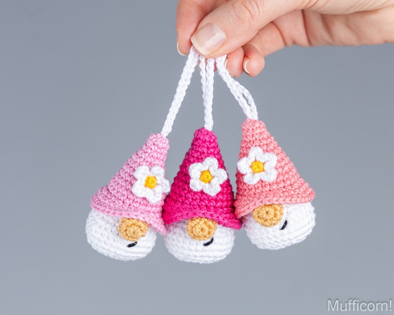 May include: Three handmade crochet gnome ornaments in shades of pink and white. Each gnome has a white face, a tan nose, and a flower on its pointed hat. The ornaments are suspended by white cords.