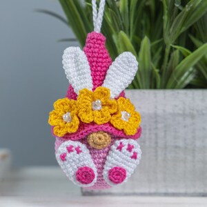 May include: A pink crocheted gnome ornament with white bunny ears and a yellow flower crown. The gnome is wearing a pink hat and has a white beard.