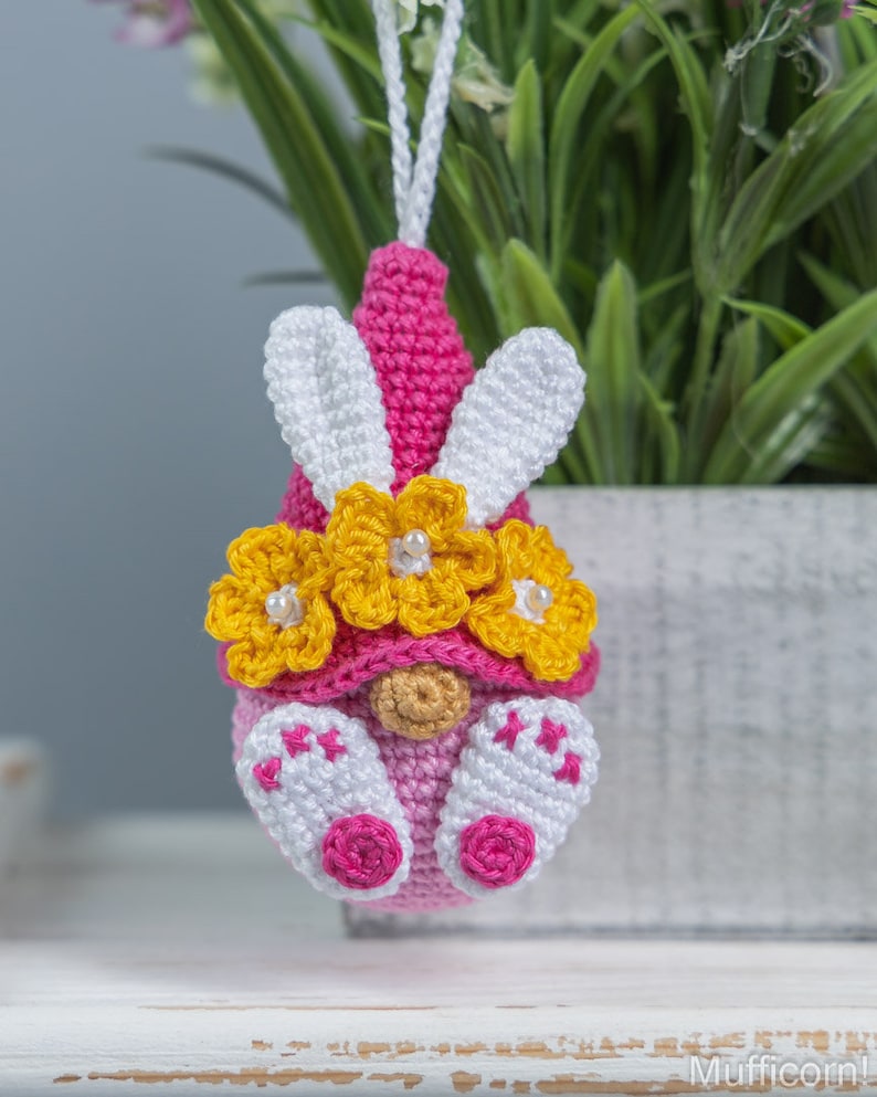 May include: A pink crocheted gnome ornament with white bunny ears and a yellow flower crown. The gnome is wearing a pink hat and has a white beard.