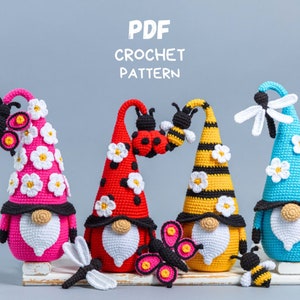May include: Four crocheted gnomes with different colored hats and insect decorations. The gnomes are wearing white beards and have black eyes. The hats are decorated with white flowers. The gnomes are sitting on a white wooden surface. The text "PDF CROCHET PATTERN" is at the top of the image.
