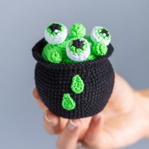 May include: A black crocheted cauldron filled with green yarn and topped with five white crocheted eyeballs with black pupils.