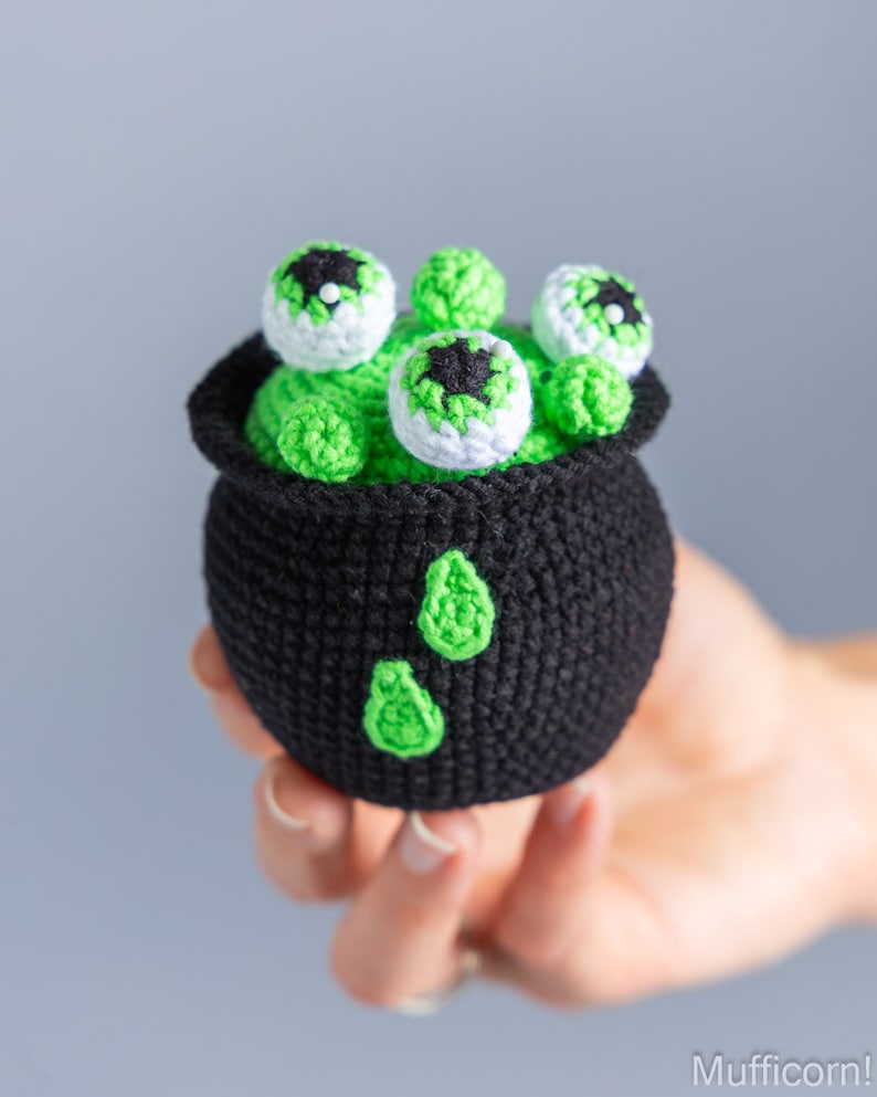 May include: A black crocheted cauldron filled with green yarn and topped with five white crocheted eyeballs with black pupils.