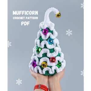 Könnte beinhalten: Ein grüner und weißer gehäkelter Weihnachtsbaum mit einem weißen gehäkelten Stern oben. Der Baum ist mit bunten Glöckchen geschmückt. Der Text "MUFFICORN CROCHET PATTERN PDF" ist oben im Bild sichtbar.