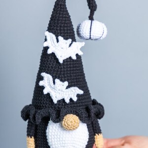 May include: A black and white crocheted gnome with a pointed hat, a white beard, and bat decorations. The gnome is holding a small white crocheted pumpkin.