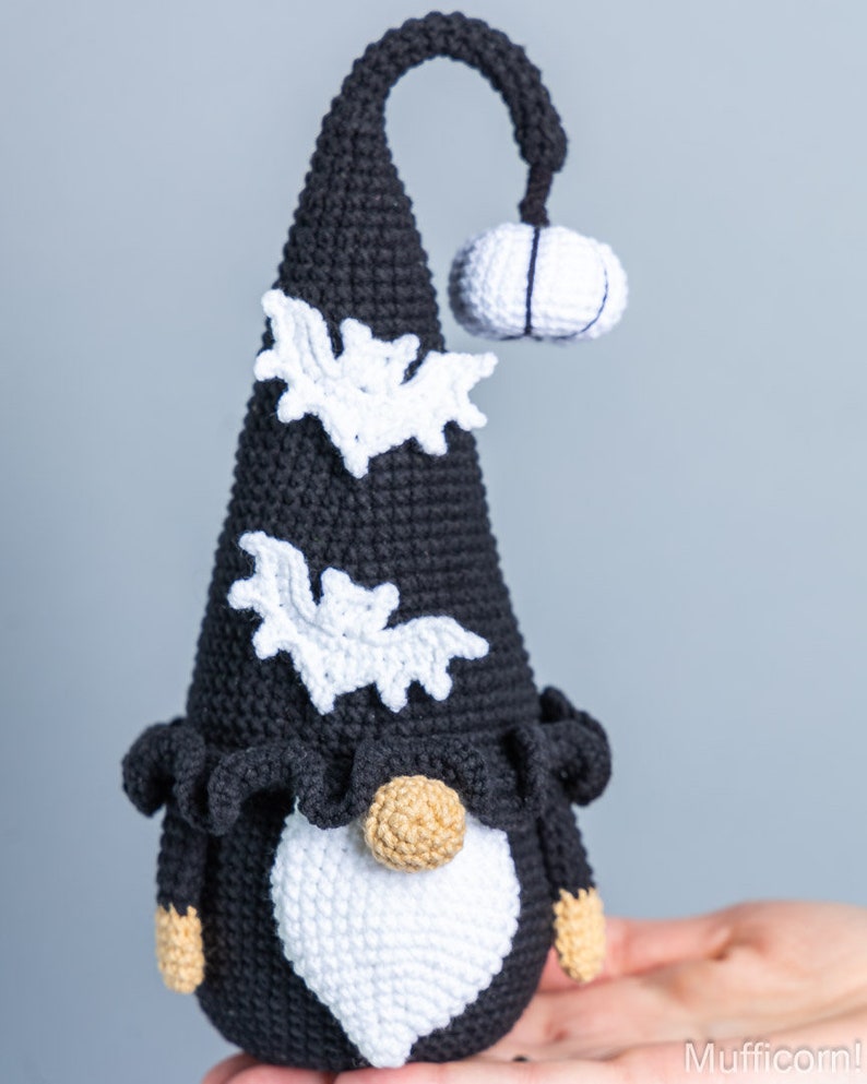 May include: A black and white crocheted gnome with a pointed hat, a white beard, and bat decorations. The gnome is holding a small white crocheted pumpkin.