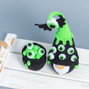 May include: A green and black crocheted gnome with a cauldron full of green goo and eyeballs. The gnome has a pointy hat with a green brim and a black brim. The gnome has a white beard and a black body. The cauldron is black with a green rim and is filled with green goo and eyeballs.