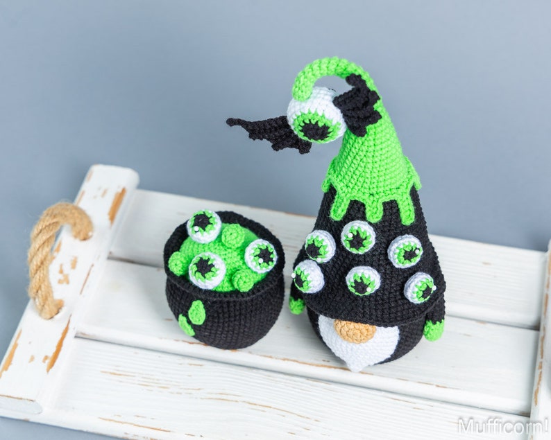 May include: A green and black crocheted gnome with a cauldron full of green goo and eyeballs. The gnome has a pointy hat with a green brim and a black brim. The gnome has a white beard and a black body. The cauldron is black with a green rim and is filled with green goo and eyeballs.