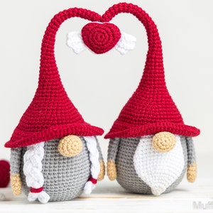 May include: Two gray crocheted gnomes with red hats and white beards are standing next to each other. Their hats are bent to form a heart shape with a small red crocheted heart with white wings in the center.