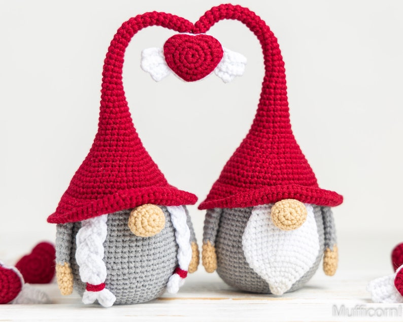 May include: Two gray crocheted gnomes with red hats and white beards are standing next to each other. Their hats are bent to form a heart shape with a small red crocheted heart with white wings in the center.