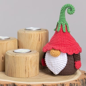 May include: A crocheted gnome with a red hat, white beard, and brown body. The gnome is sitting on a wooden log with three candle holders.
