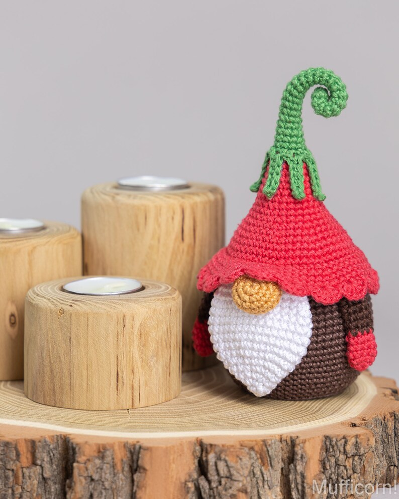 May include: A crocheted gnome with a red hat, white beard, and brown body. The gnome is sitting on a wooden log with three candle holders.