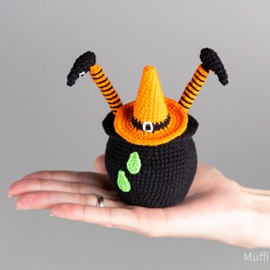 May include: A crocheted witch cauldron with orange and black striped legs and a pointed orange hat. The cauldron is black with green accents.