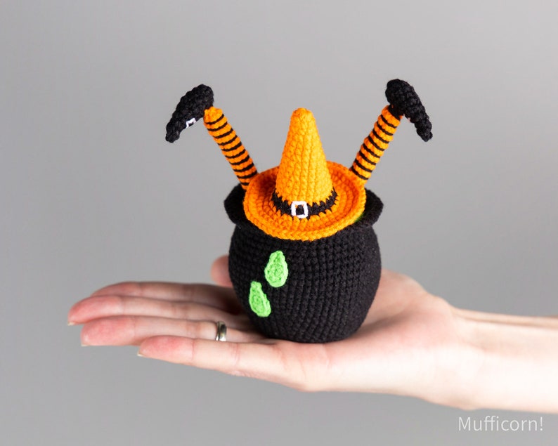 May include: A crocheted witch cauldron with orange and black striped legs and a pointed orange hat. The cauldron is black with green accents.