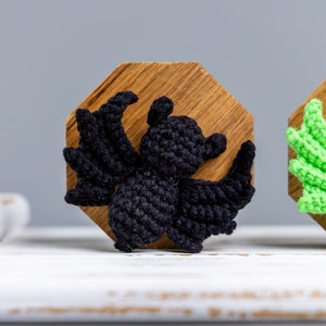 May include: A black crocheted bat figurine with wings spread wide, displayed on a wooden octagon-shaped base.