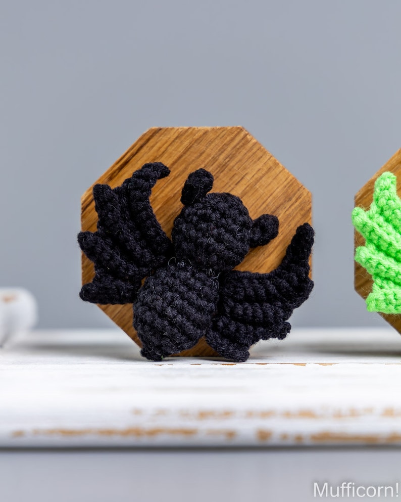 May include: A black crocheted bat figurine with wings spread wide, displayed on a wooden octagon-shaped base.