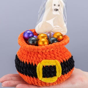 May include: A crocheted orange and black cauldron-shaped basket with a yellow buckle detail, filled with assorted foil-wrapped chocolate candies and a single ghost-shaped lollipop in a clear plastic wrapper, featuring the text Mufficorn!.
