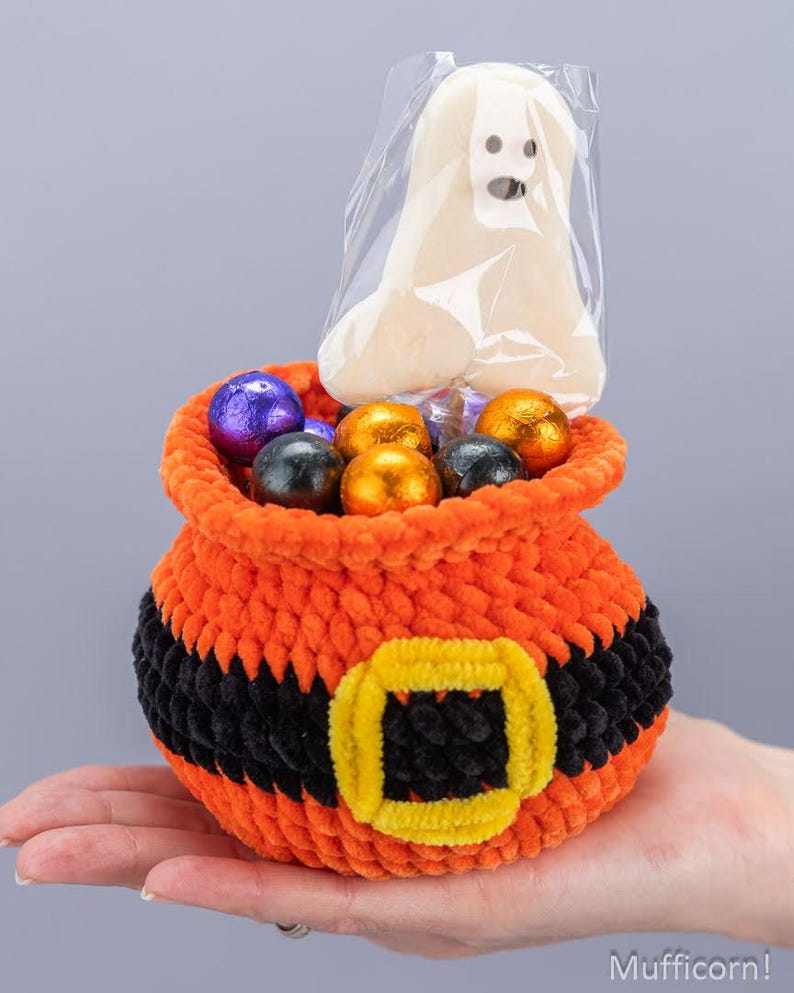 May include: A crocheted orange and black cauldron-shaped basket with a yellow buckle detail, filled with assorted foil-wrapped chocolate candies and a single ghost-shaped lollipop in a clear plastic wrapper, featuring the text Mufficorn!.