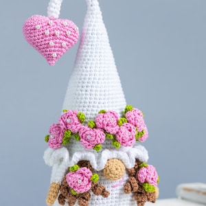 May include: White crocheted gnome with a pink heart on top of its hat. The gnome has a pink rose crown and pink roses on its face. The gnome is sitting on a white surface.