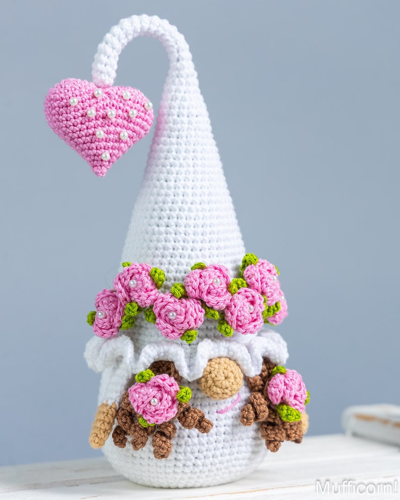 May include: White crocheted gnome with a pink heart on top of its hat. The gnome has a pink rose crown and pink roses on its face. The gnome is sitting on a white surface.