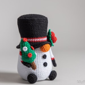 May include: A crocheted snowman wearing a black top hat with a red and white band, holding a green Christmas tree in one hand. The snowman has orange carrot nose, black eyes, and black buttons.