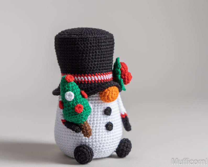 May include: A crocheted snowman wearing a black top hat with a red and white band, holding a green Christmas tree in one hand. The snowman has orange carrot nose, black eyes, and black buttons.