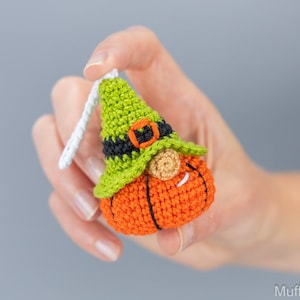 May include: A crocheted pumpkin gnome ornament with a green hat, black belt, and white beard. The gnome is holding a small white loop for hanging.