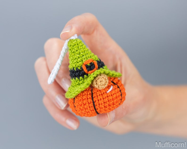May include: A crocheted pumpkin gnome ornament with a green hat, black belt, and white beard. The gnome is holding a small white loop for hanging.