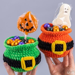 May include: Two crocheted baskets shaped like cauldrons, one green and one orange, each featuring a black band with a yellow buckle. The green basket holds a pumpkin-shaped lollipop, and the orange basket holds a ghost-shaped treat in plastic.