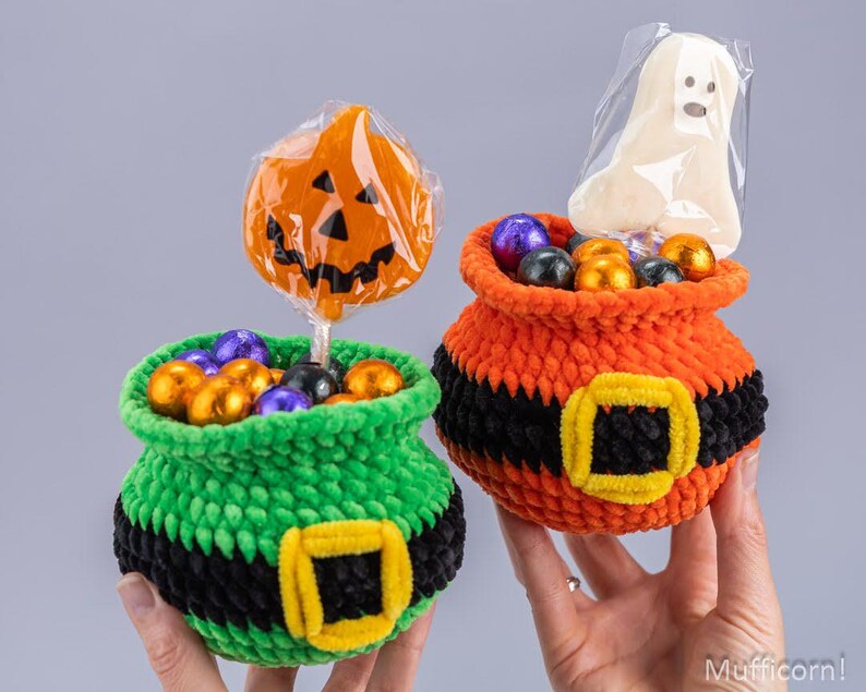 May include: Two crocheted baskets shaped like cauldrons, one green and one orange, each featuring a black band with a yellow buckle. The green basket holds a pumpkin-shaped lollipop, and the orange basket holds a ghost-shaped treat in plastic.
