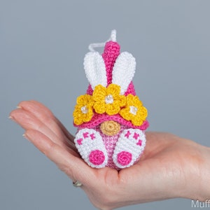 May include: A pink crocheted gnome with white bunny ears and a yellow flower crown. The gnome is holding a small pink heart in its hand.