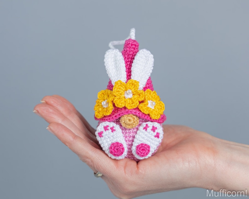 May include: A pink crocheted gnome with white bunny ears and a yellow flower crown. The gnome is holding a small pink heart in its hand.