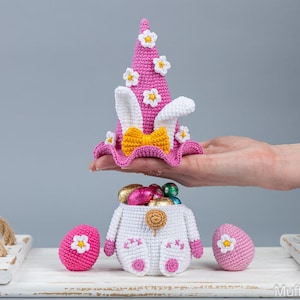 May include: A crocheted Easter bunny basket with pink and white ears, a pink hat with white flowers, and a yellow bow. The basket is filled with chocolate eggs and is sitting on a white wooden tray.
