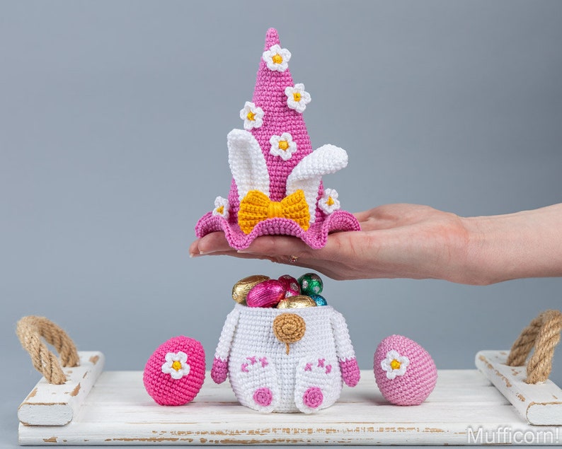 May include: A crocheted Easter bunny basket with pink and white ears, a pink hat with white flowers, and a yellow bow. The basket is filled with chocolate eggs and is sitting on a white wooden tray.