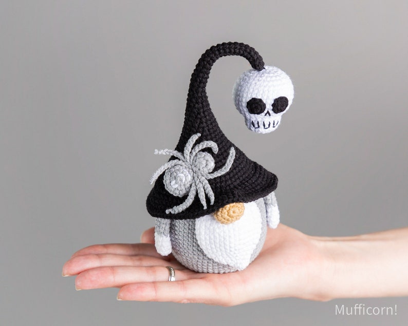 May include: A gray and white crocheted gnome wearing a black witch hat with a spider and a white skull on top.