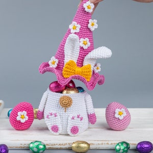 May include: A crocheted Easter bunny basket with a pink hat and a yellow bow. The basket is filled with chocolate eggs and there are more eggs scattered around it.